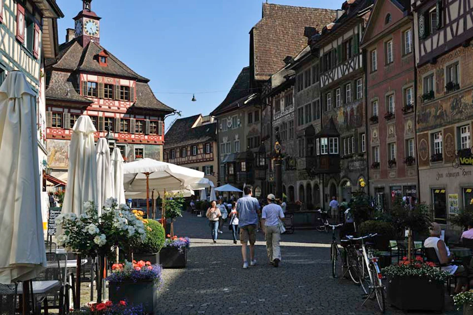 People walking on a cobblestone street with historic buildings, an outdoor café with umbrellas, potted flowers, and bicycles.