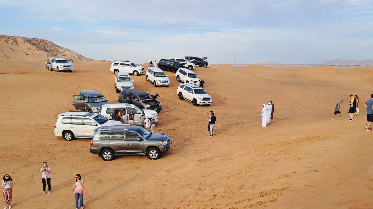 SUVs estacionados em dunas de areia, com pessoas por perto, algumas vestindo trajes tradicionais brancos e outras com roupas casuais.