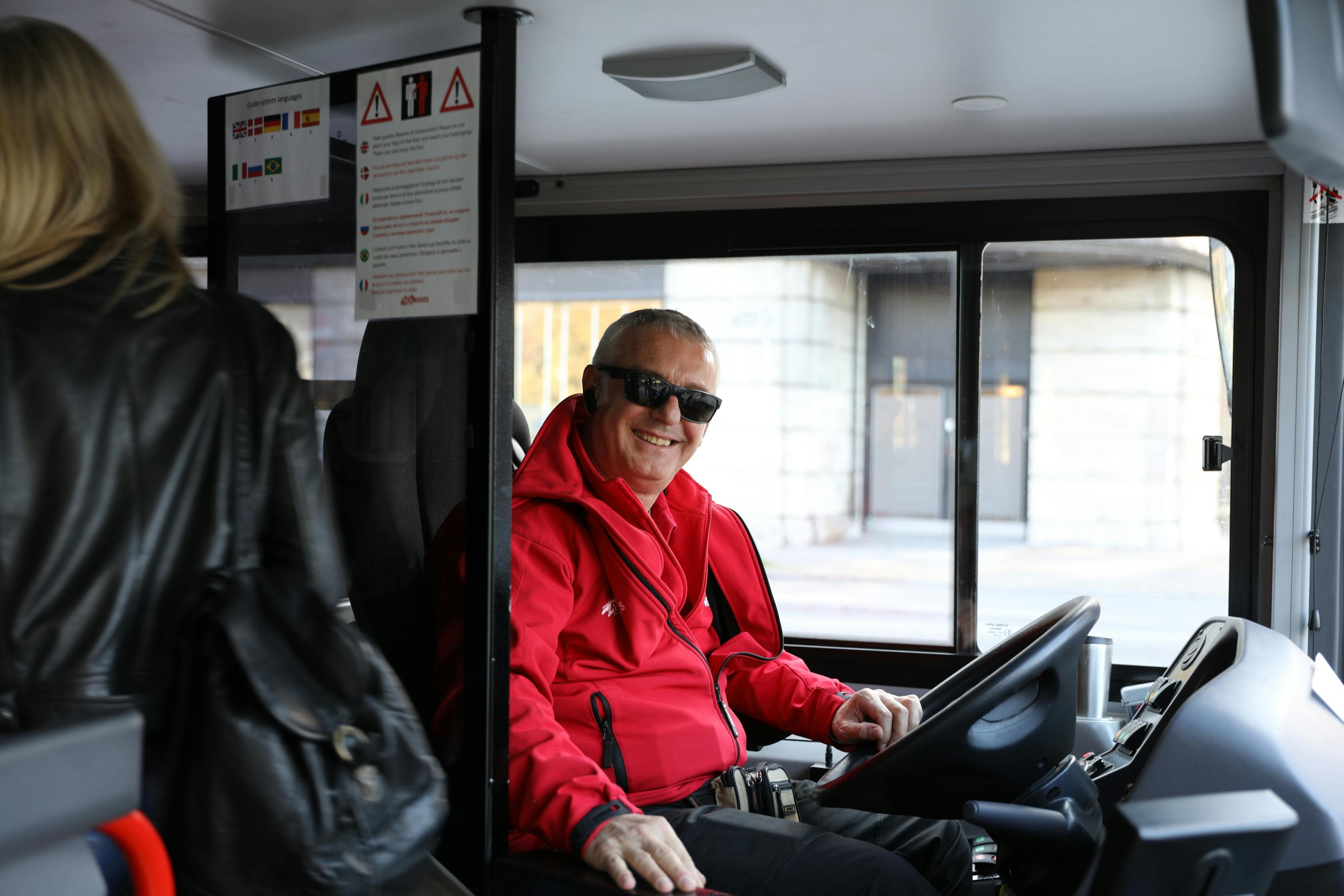 A man in a red jacket and sunglasses smiling while sitting in the driver's seat of a bus. Signs with multiple flags are above him.