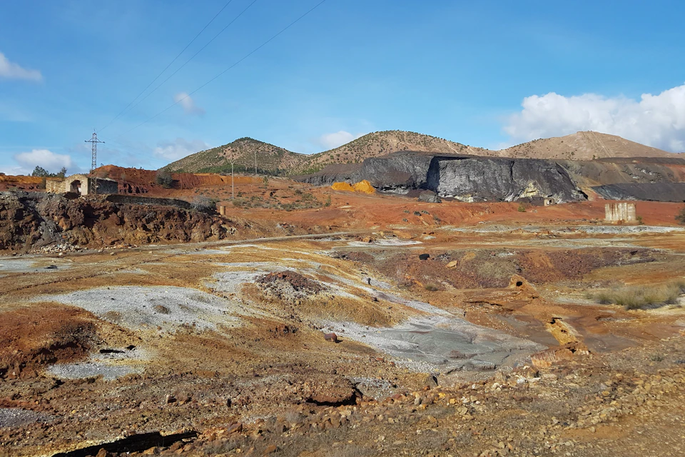 A barren, rocky landscape with multi-colored earth and distant hills under a blue sky with scattered clouds.