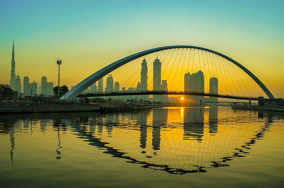 Arch bridge spanning over calm water with city skyscrapers in the background at sunset, reflecting golden hues.