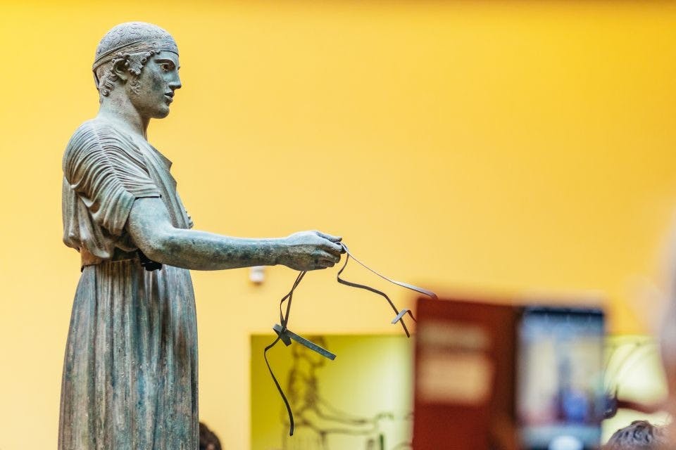 노란색 실내 박물관을 배경으로 얇고 섬세한 물건을 들고 있는 서 있는 모습의 고대 청동 동상.