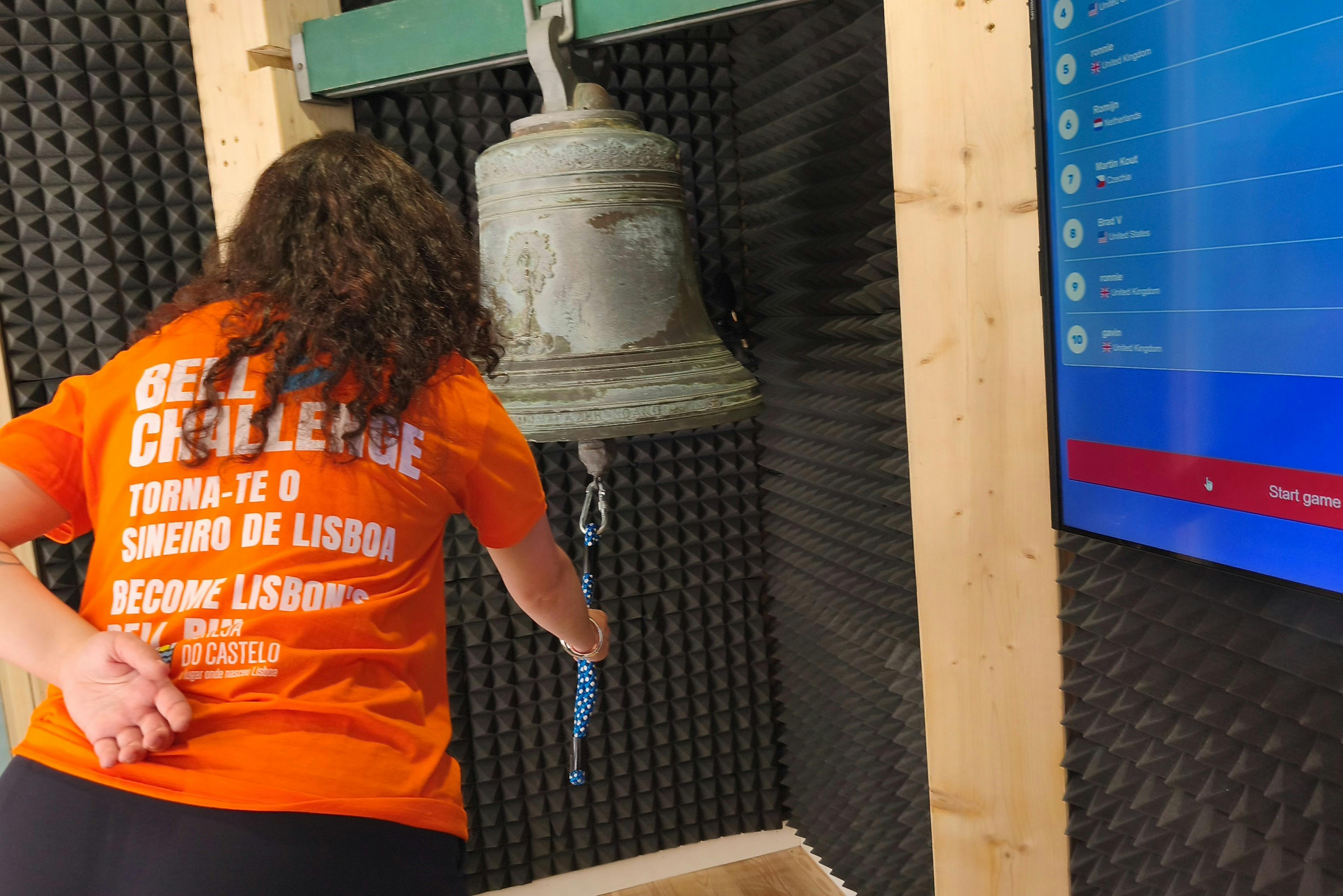 A person wearing an orange shirt rings a large bell in a soundproofed room. A digital display is visible on the right.