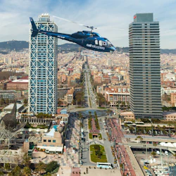 A blue helicopter flies over a busy cityscape with tall buildings, a long central avenue, and pedestrian areas below.
