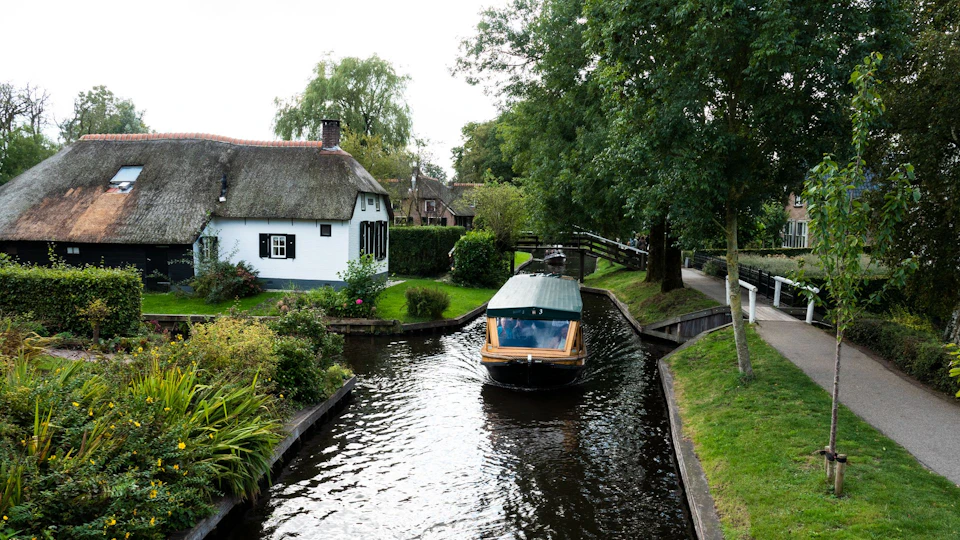 A boat travels along a narrow canal surrounded by green vegetation and houses with thatched roofs.