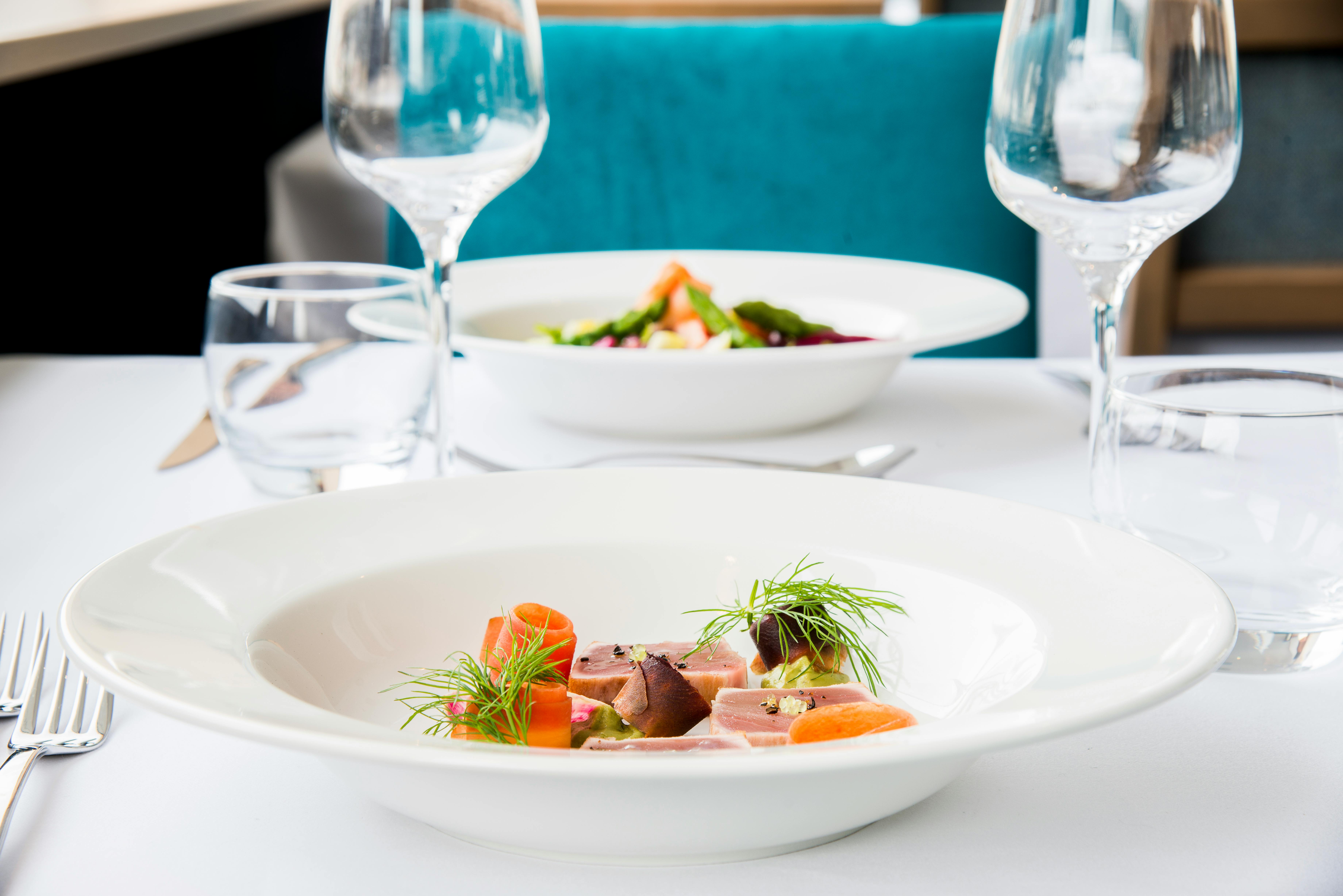 Elegant borddækning med to tallerkener med gourmetmad, vinglas og en turkis stol i baggrunden.