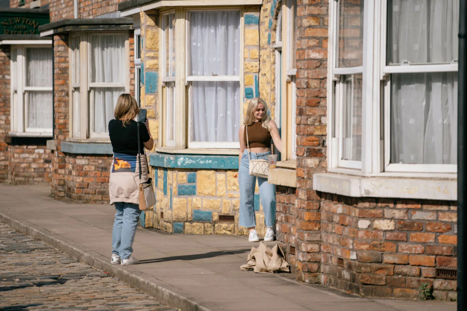 Two women on a sunlit sidewalk; one taking a photo with a phone, the other posing against a brick and stone wall.