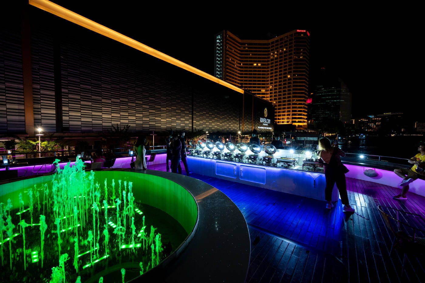 화려한 분수, 사람들이 어우러지고 도시의 스카이라인을 배경으로 일련의 조명이 있는 나이트클럽 옥상 풍경.