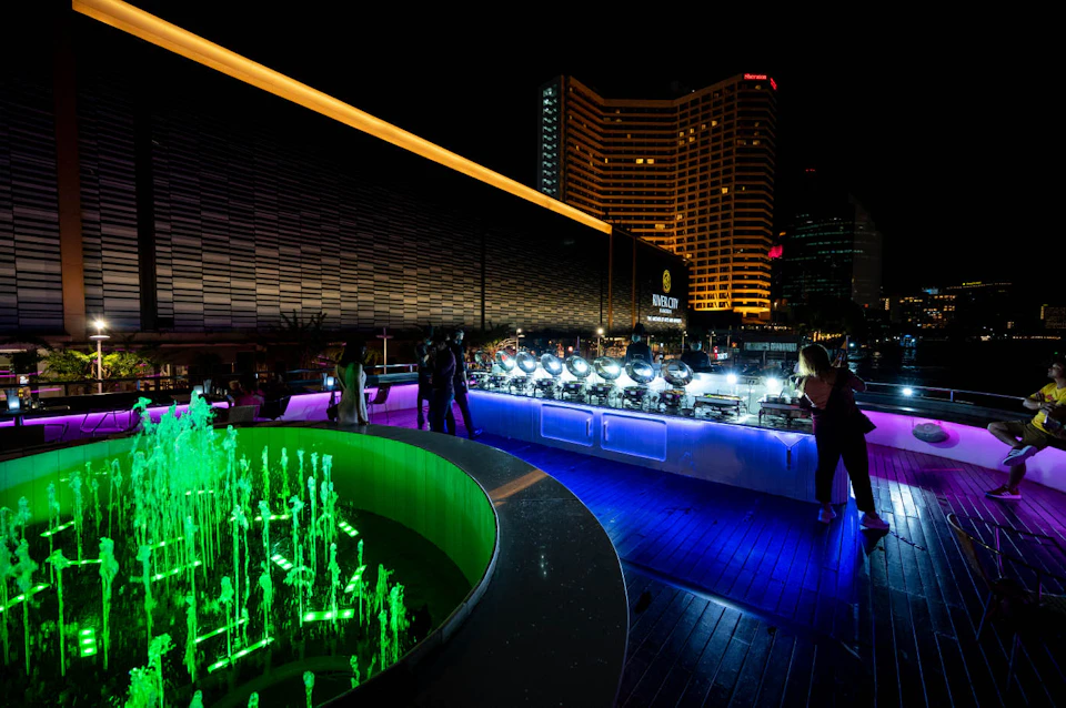 Nightclub rooftop scene with colorful illuminated fountains, people mingling, and a row of lights against a city skyline backdrop.
