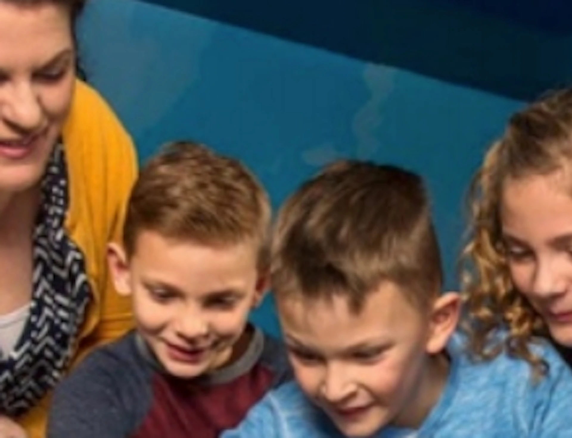 Three children and a woman smile and point at marine life in a touch tank at an aquarium. Bright coral and fish are visible.
