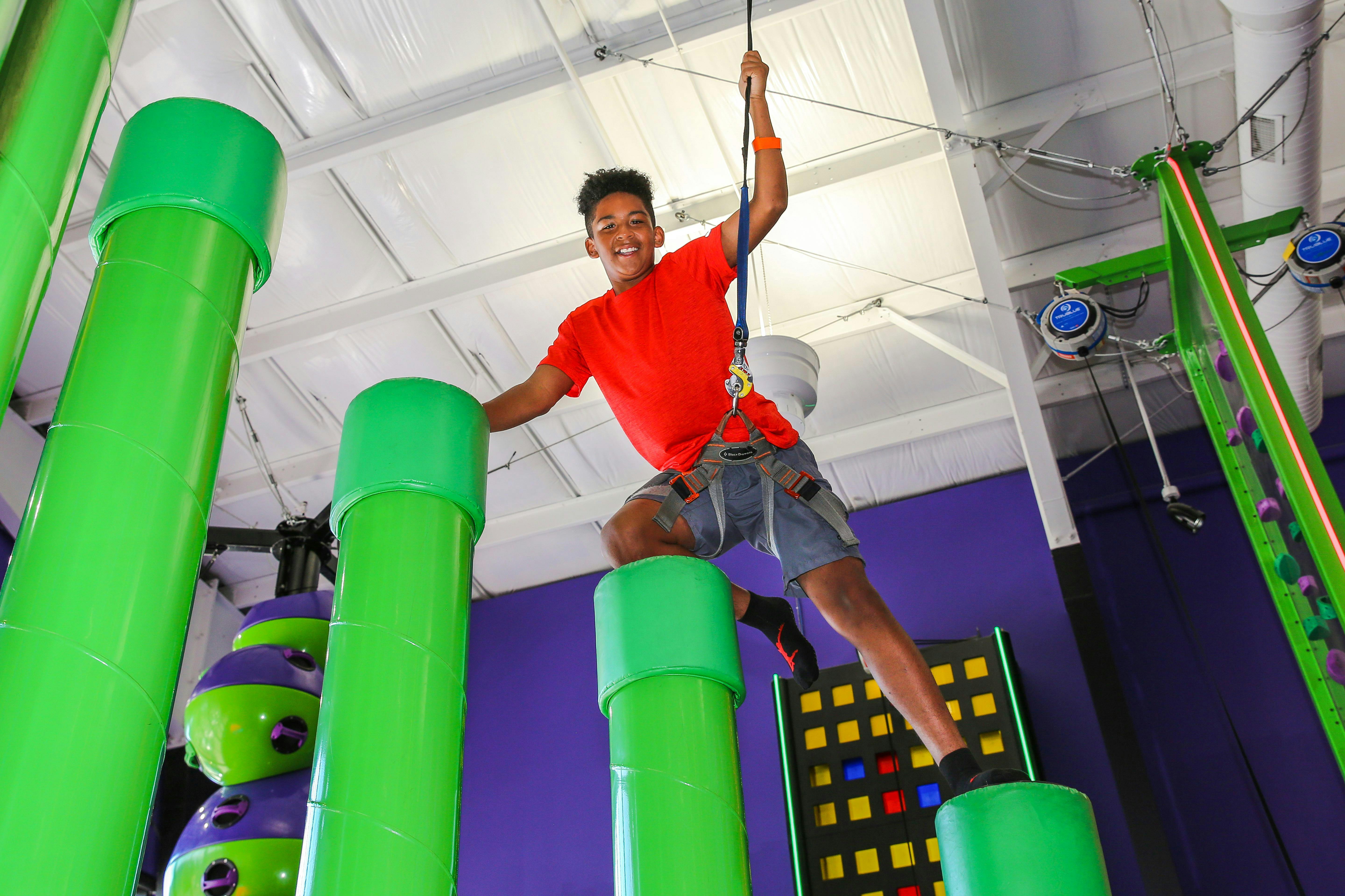 A boy in a red shirt climbs green cylindrical structures indoors, connected to a safety harness. He smiles while holding a rope.
