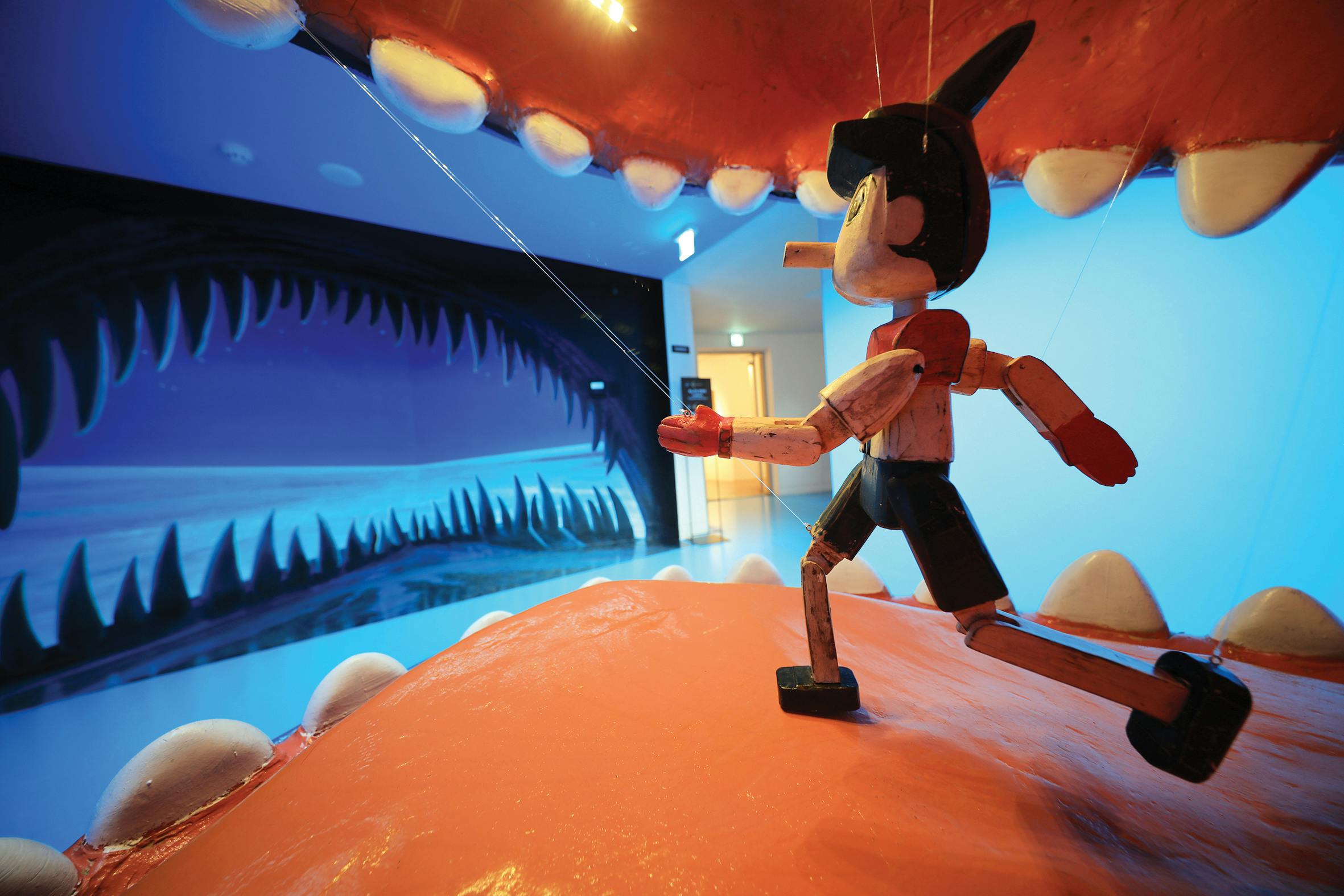 A wooden puppet resembling Pinocchio inside a large mouth, with a painted background depicting a sea and shark teeth.