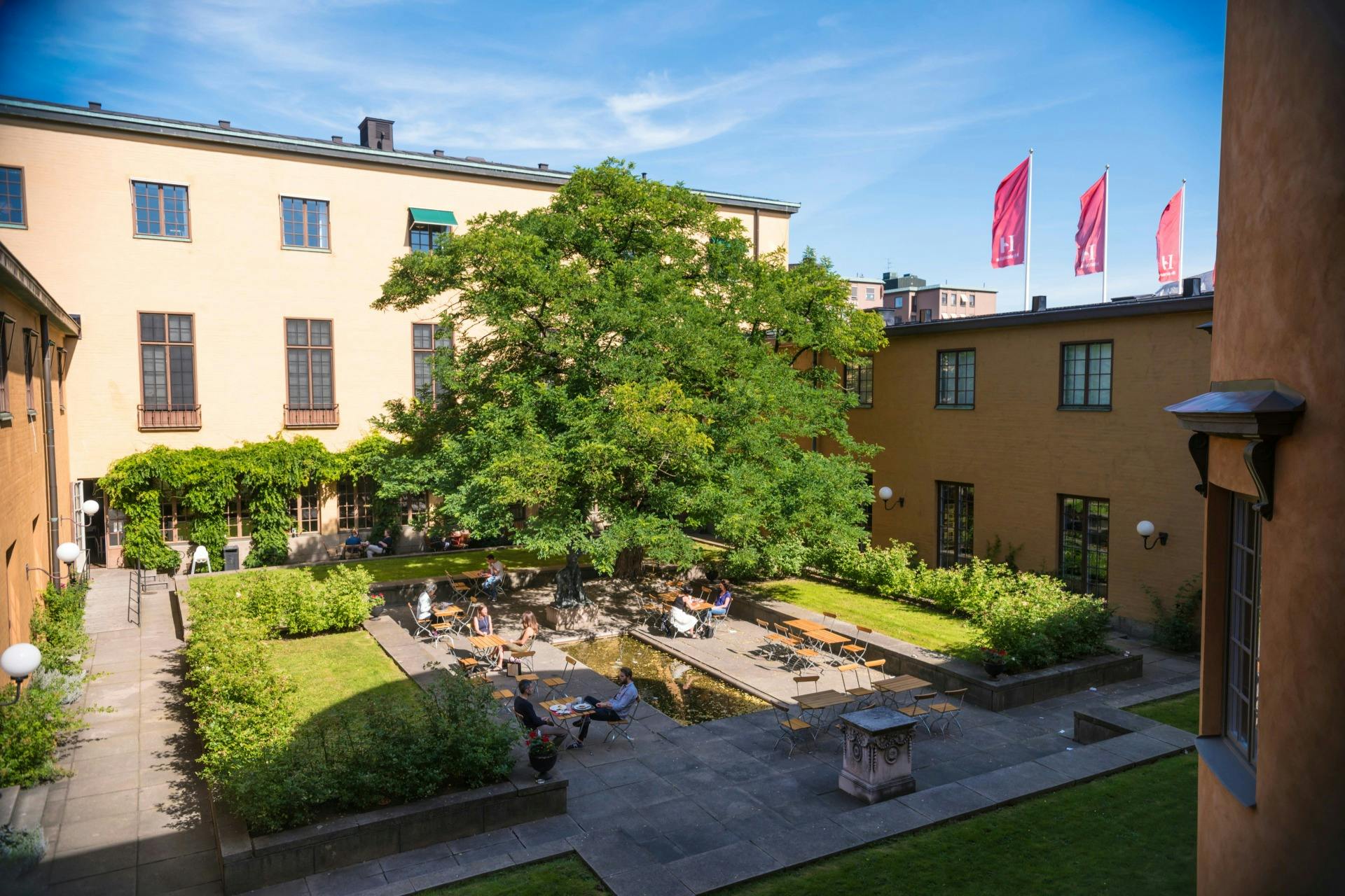 En innergård med ett träd, människor som sitter vid bord, omgivna av gula byggnader under en blå himmel med röda flaggor synliga.