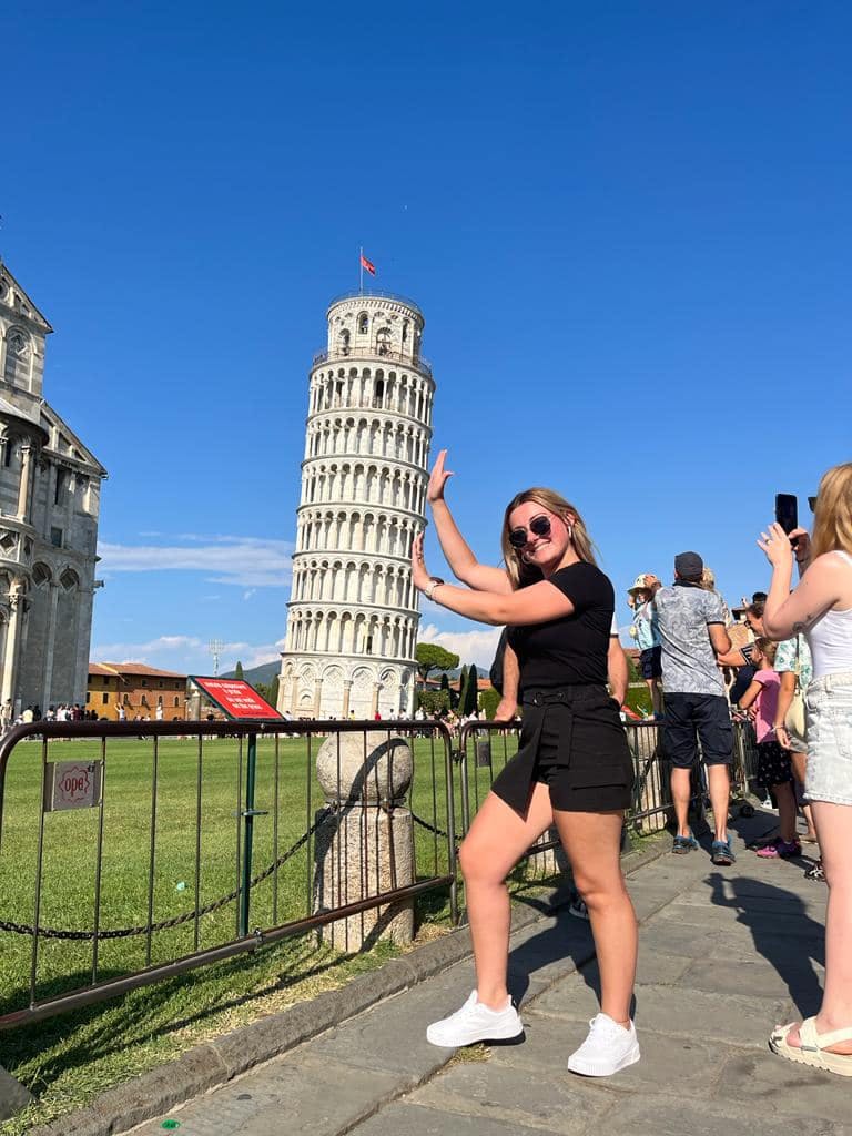 A woman playfully poses as if holding up the Leaning Tower of Pisa, with a clear blue sky and other tourists in the background.