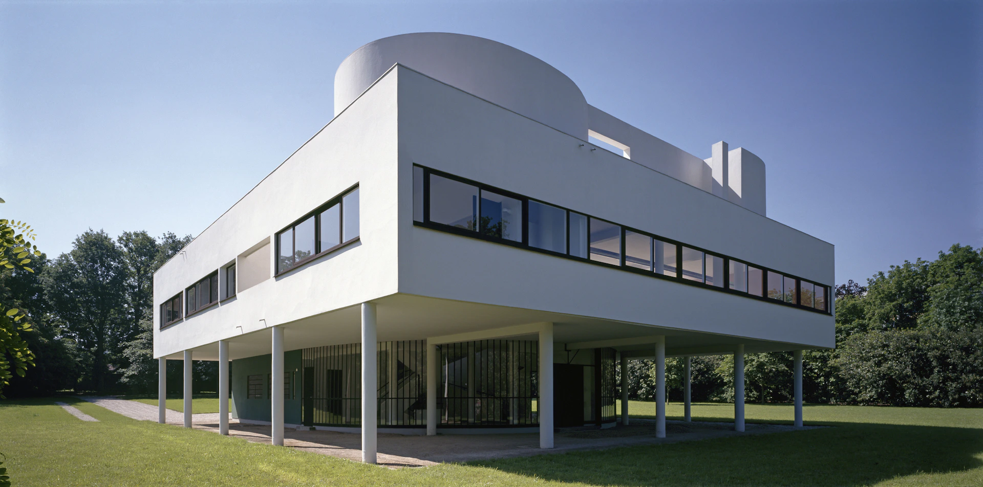 Modernist white building with large rectangular windows, supported by thin columns, set on a grassy lawn under a clear sky.