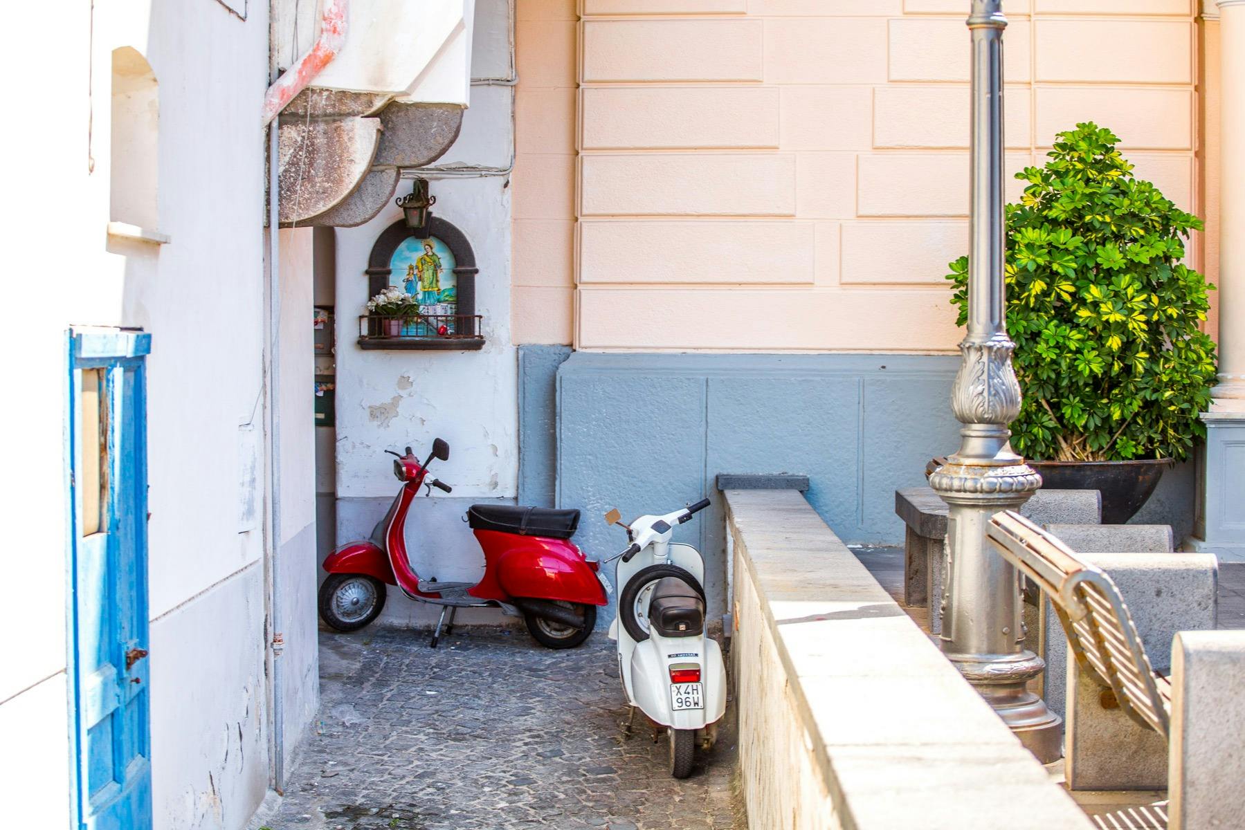 Twee scooters, een rode en een witte, parkeerden in een steegje naast een perzikkleurige muur met een religieus heiligdom erop.