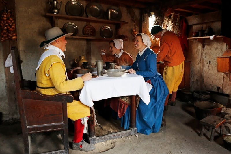 Lidé oblečení v historických šatech sedí a stojí kolem stolu v rustikální místnosti s dřevěnými policemi a kuchyňským náčiním.