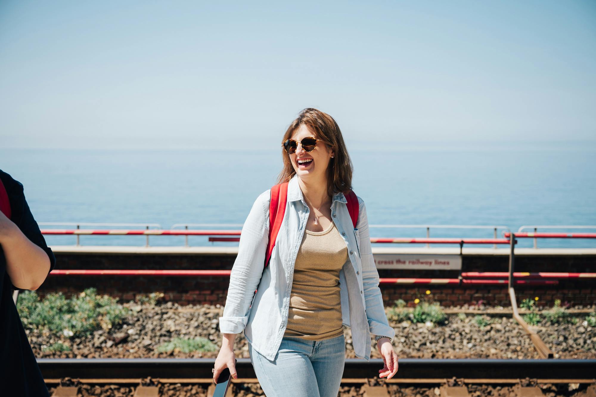 一位戴着太阳镜、背着红色背包的女士走在火车轨道附近，背景中可以看到大海。