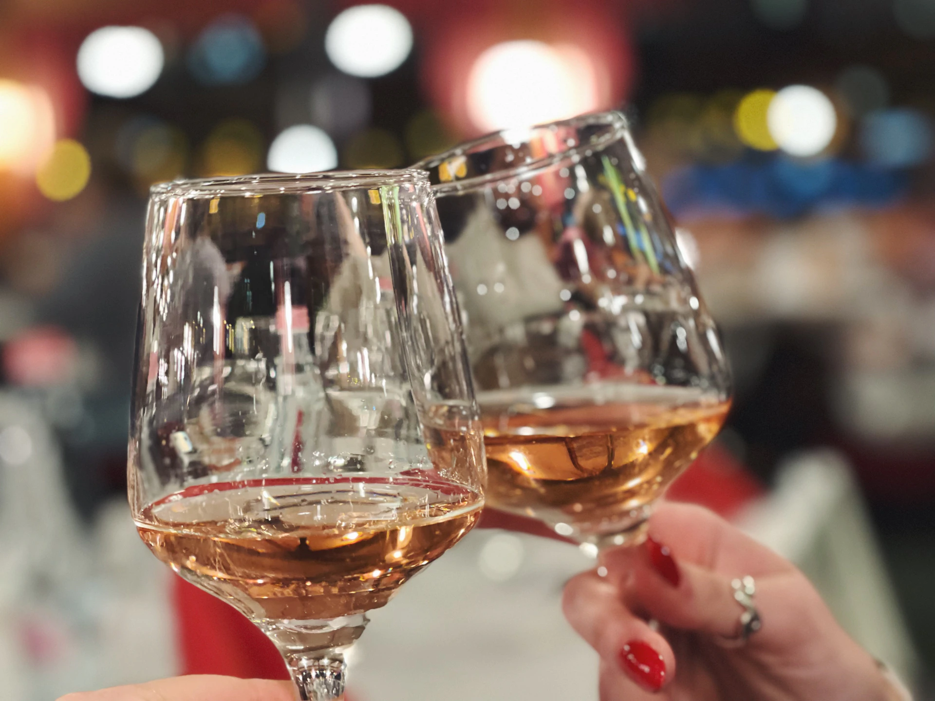 Two wine glasses clink in a toast, held by hands with red-painted nails, against a blurred background with colorful lights.