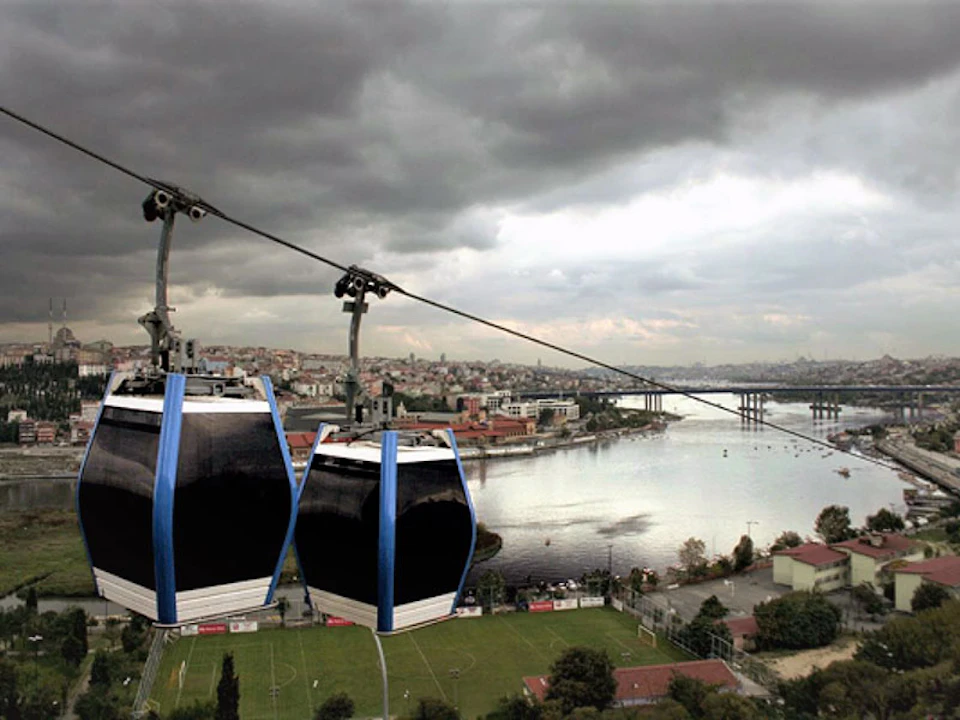 Two gondola lifts suspended over a river with a cityscape in the background under a cloudy sky.