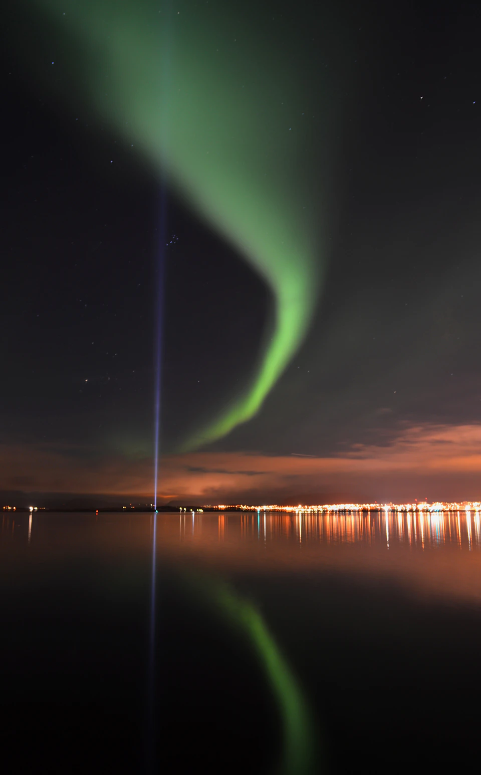 Green aurora borealis over a night sky with a vertical blue light beam near a cityscape reflected on the water.