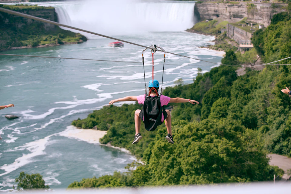 Person zip-lining over a river with a waterfall in the background, surrounded by dense greenery.