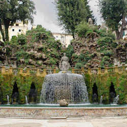 En stenfontän med vatten som strömmar från valv under statyer omgivna av mossbeklädda klippor och frodig grönska.