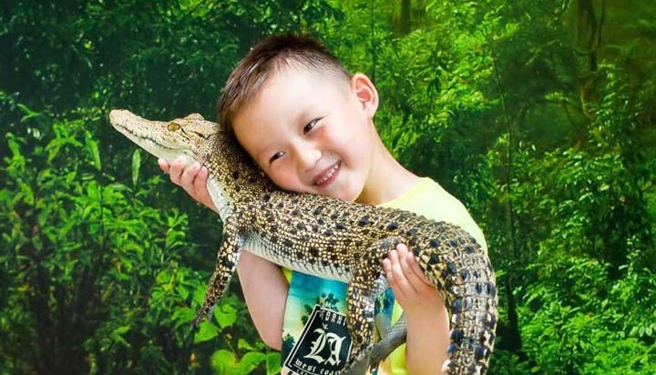 一个微笑的孩子把一只小鳄鱼抱在脸颊上，站在郁郁葱葱的绿色丛林背景前。