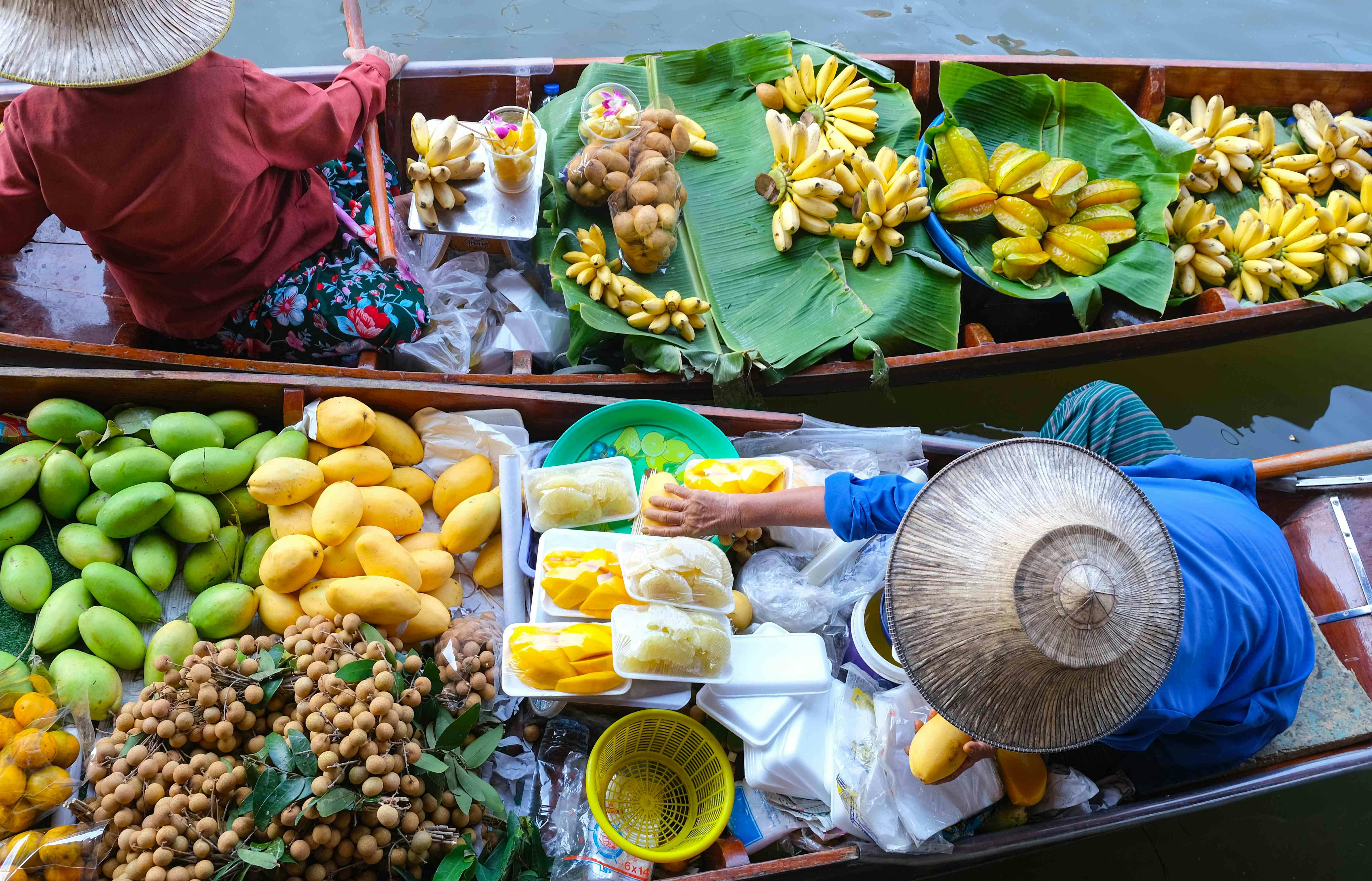 Two boats at a floating market filled with tropical fruits, including bananas, mangoes, and longans, arranged on banana leaves.