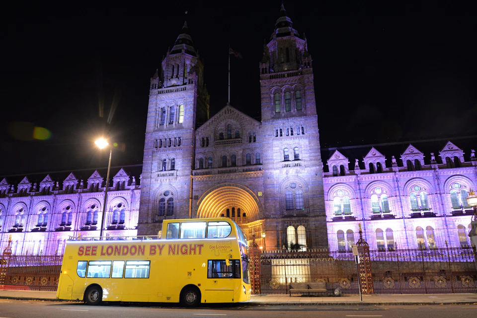 A yellow double-decker bus reading 'See London by Night' parked in front of a lit historic building at night.