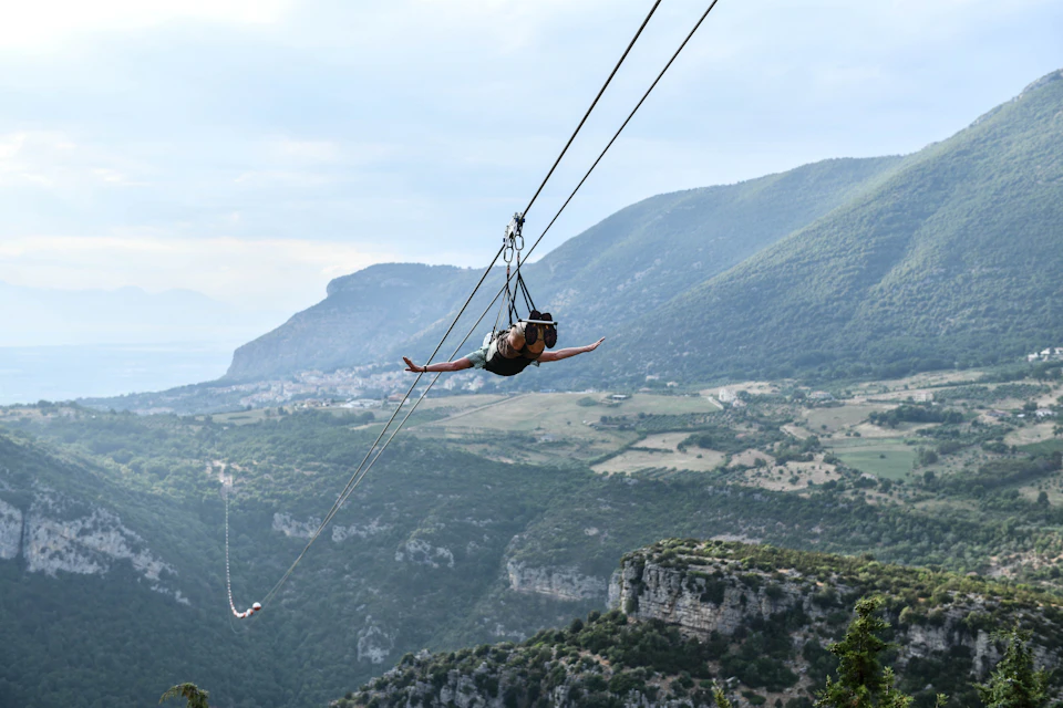 Person zip-lining over a scenic mountainous landscape with valleys and greenery under a partly cloudy sky.