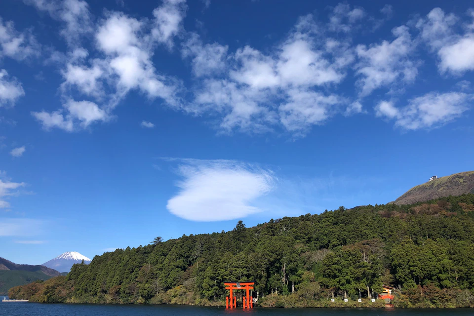 A red torii gate stands in the water near a forested shoreline under a blue sky with scattered clouds.