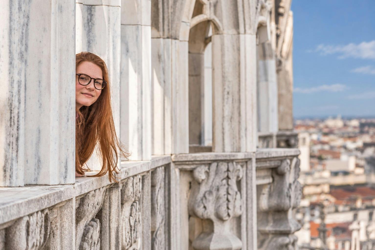 Žena s dlouhými zrzavými vlasy a brýlemi vykukující z mramorové architektonické struktury s vyřezávanými detaily.