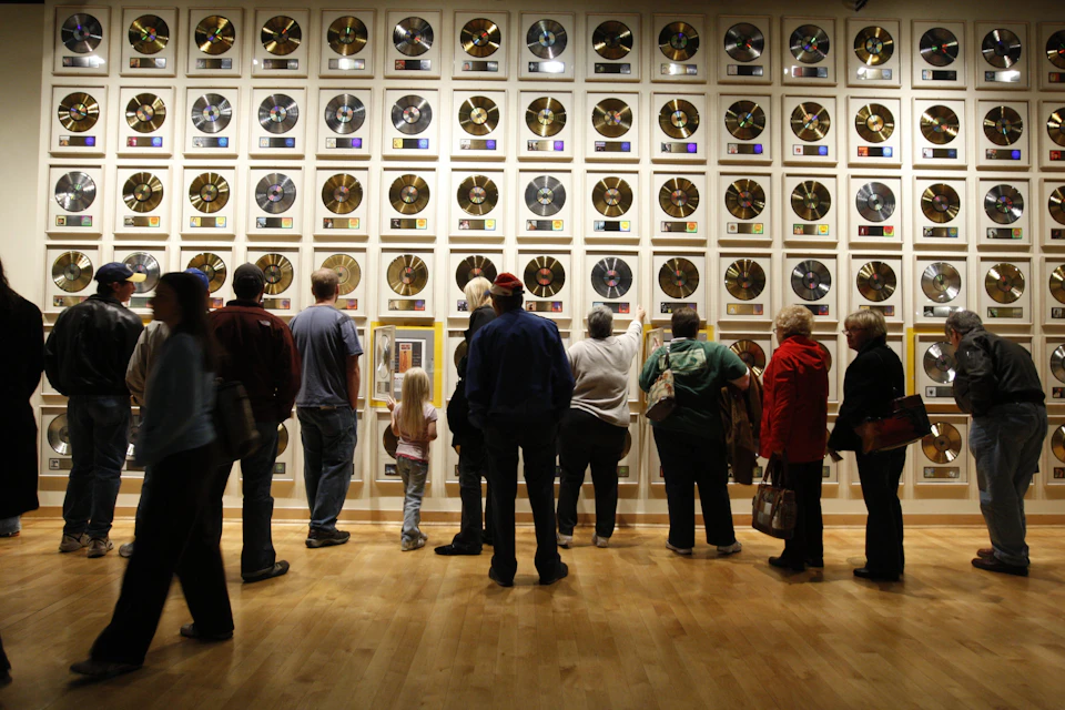 People viewing a large wall of framed gold and platinum records in a museum or gallery setting on a wooden floor.