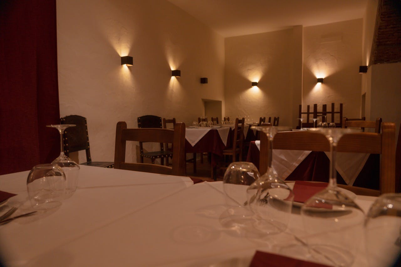 Špatně osvětlený interiér restaurace s několika prázdnými stoly s bílými ubrusy, sklenicemi na víno a červenými ubrousky.