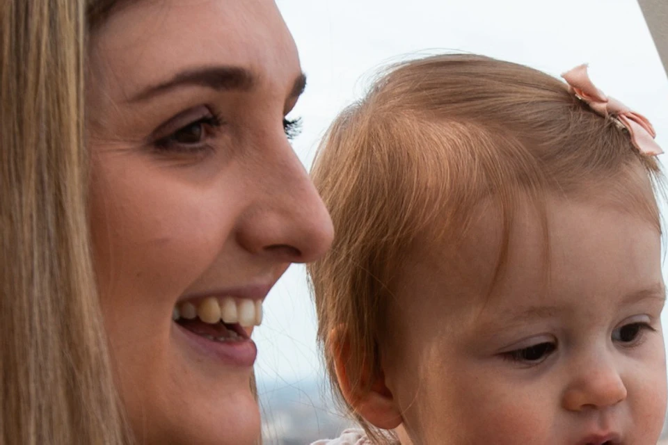 A woman holding a baby inside a large observation wheel pod, smiling and looking out at the cityscape below.