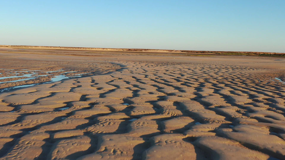 Sandy beach at low tide showing rippled patterns with a clear blue sky in the background.