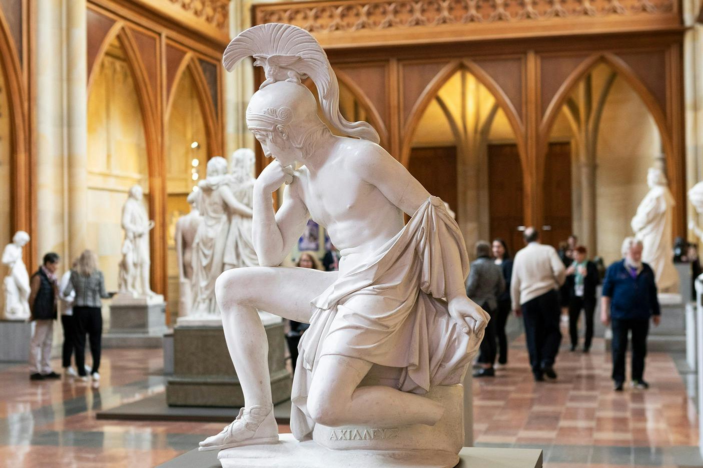 A marble statue in a museum setting, depicting a seated figure with a helmet, surrounded by other sculptures and visitors.
