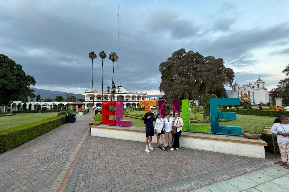 Four people pose in front of colorful large letters spelling "ELUE" in a paved plaza with trees and a building in the background.