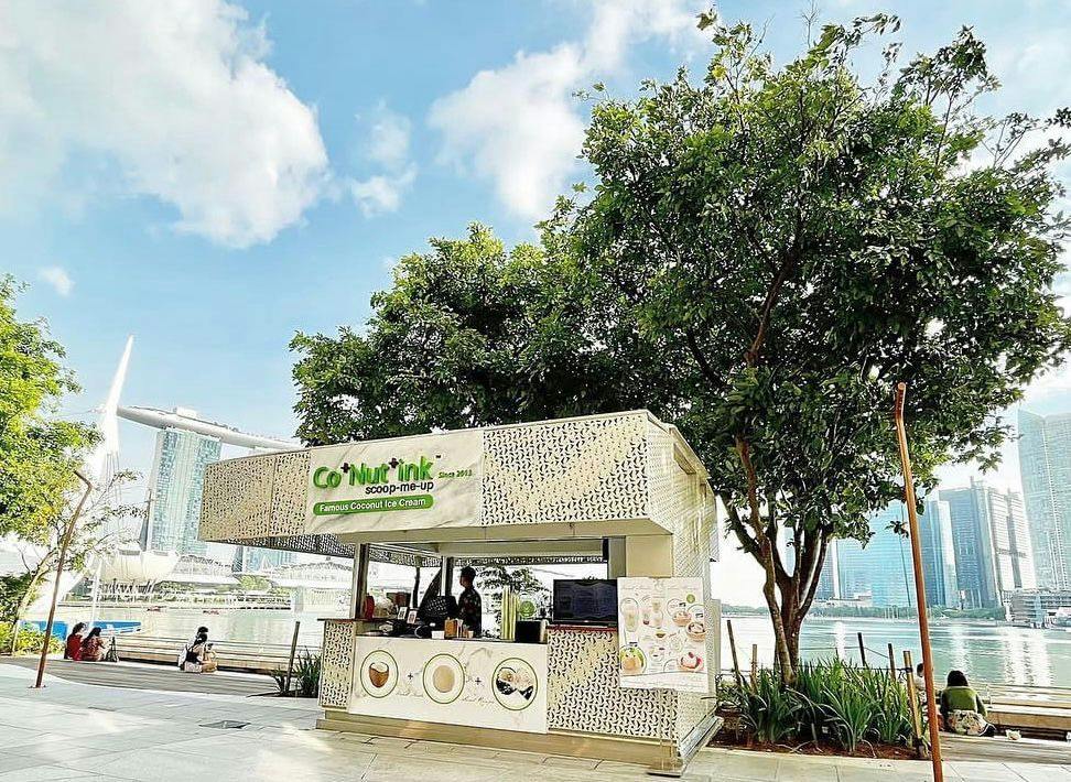 An outdoor ice cream stand named 'Co+Nut+ink' is set up under a tree, with a cityscape and waterfront visible in the background.
