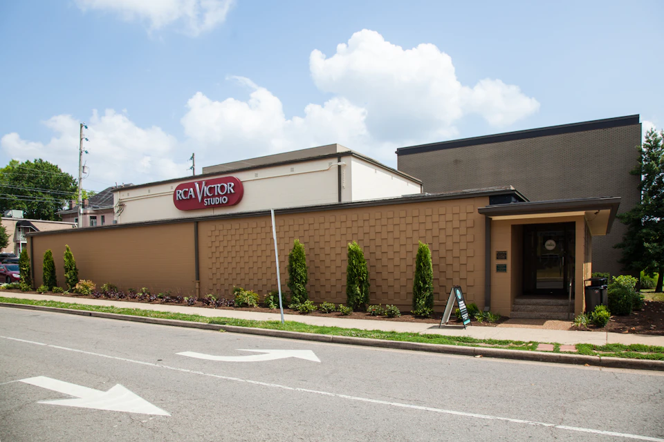 A beige building with a sign reading "RCA Victor Studio," a sidewalk, and small bushes, under a blue sky with clouds.