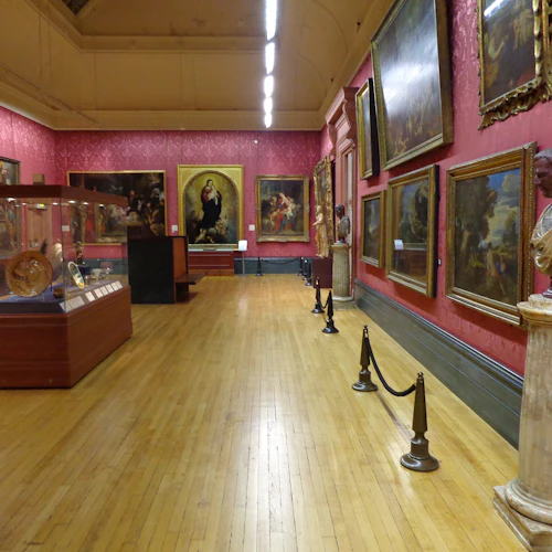 Konstgalleri med röda väggar, trägolv, olika målningar och skulpturer. Centralt glasfodral visar dekorativa föremål.