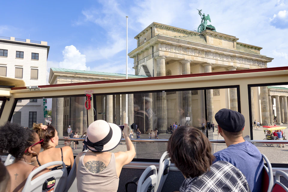 Tourists on an open-top bus view the Brandenburg Gate in Berlin on a sunny day, with people walking nearby.