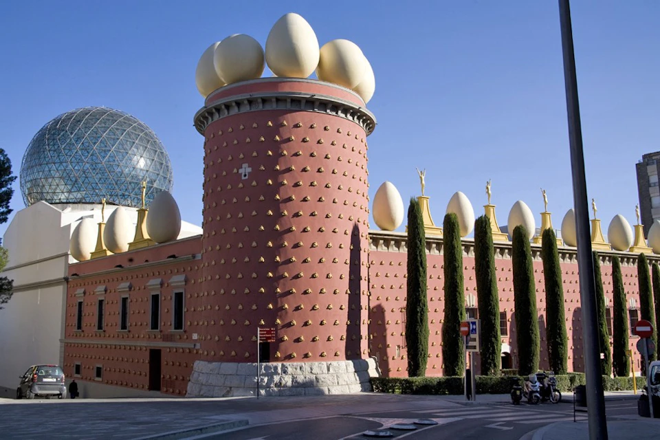 A red cylindrical building with small golden sculptures and large eggs atop, adjacent to tall cylindrical columns with greenery.