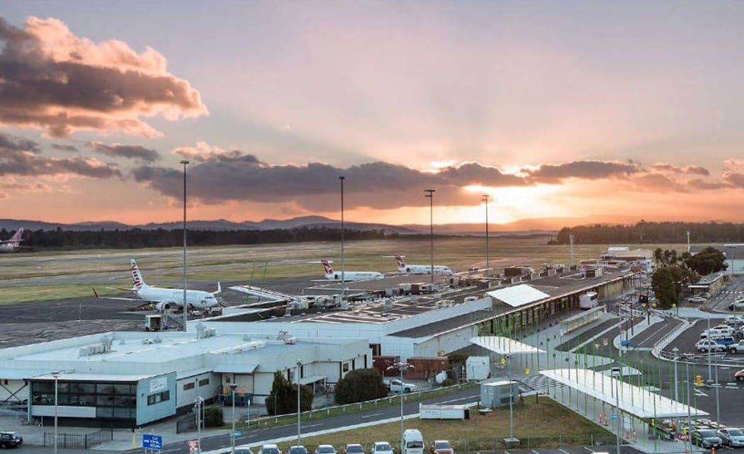 Un aéroport au coucher du soleil avec des avions garés aux portes d'embarquement, un terminal et des montagnes lointaines sous un ciel partiellement nuageux.