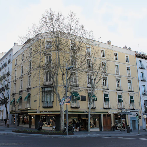 En stadsgata med en hörnbyggnad med bostadsbutiker, med flera våningar med balkonger, träd framför och fotgängare.