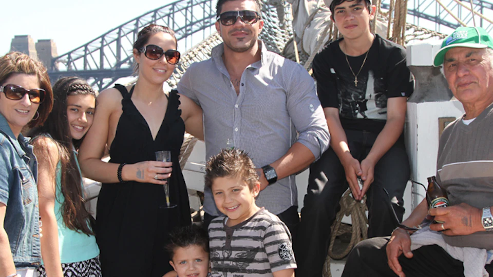 A group of seven people smiling and posing on a boat with a bridge visible in the background on a sunny day.