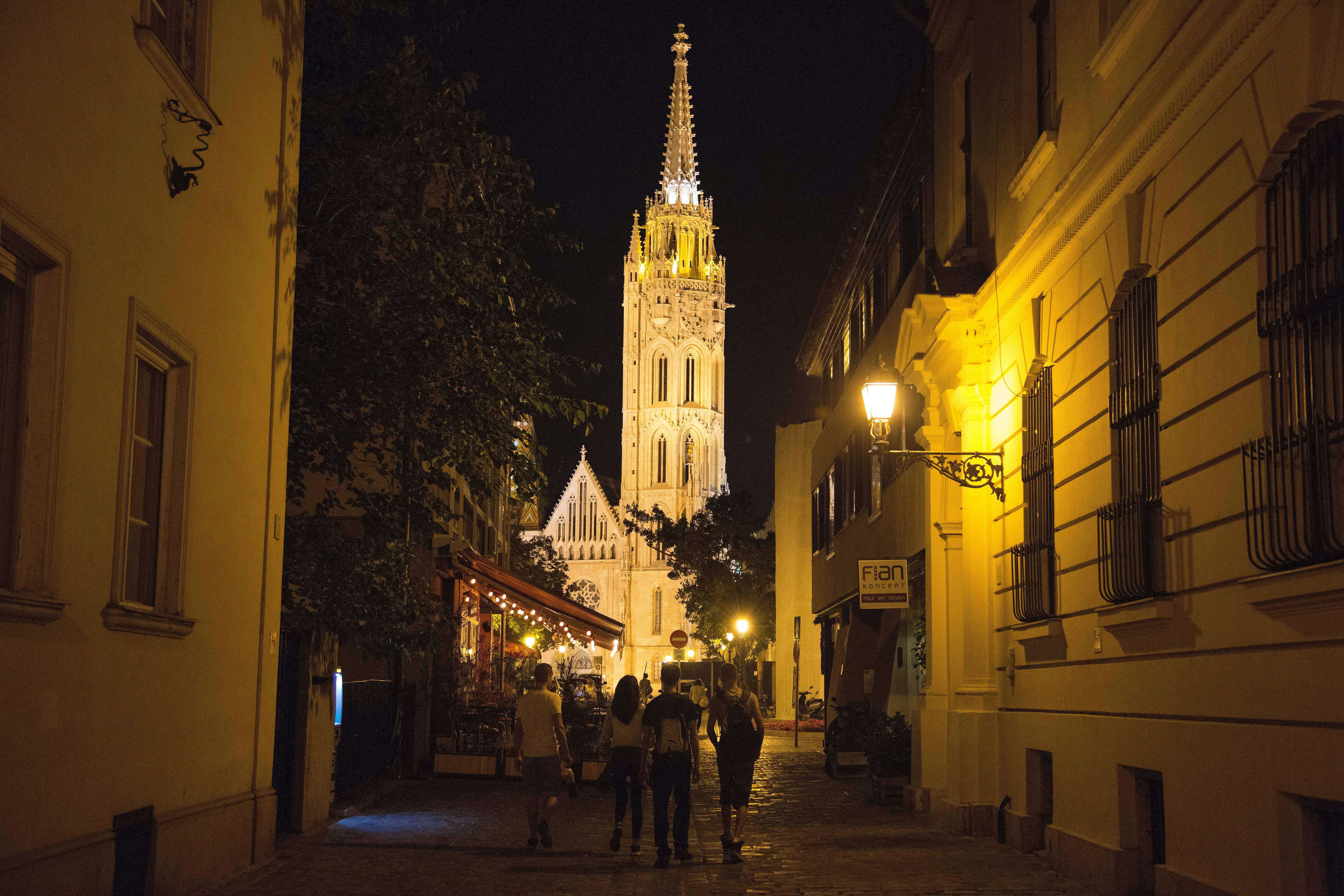 Four people walk through a dimly lit street towards a tall, illuminated Gothic-style church tower at night.