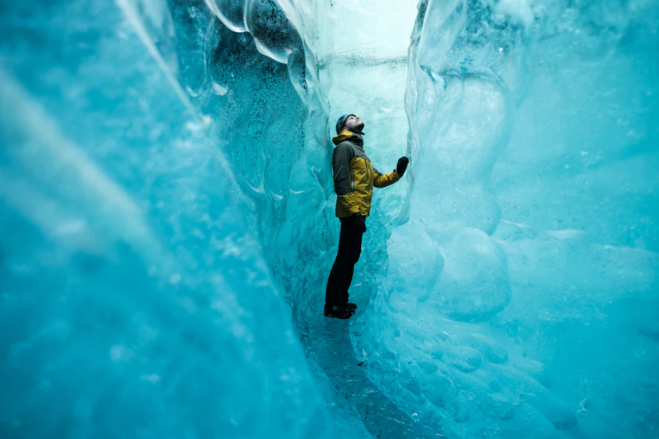 A person in a yellow jacket stands between towering blue ice walls, looking upward in a narrow ice cave.