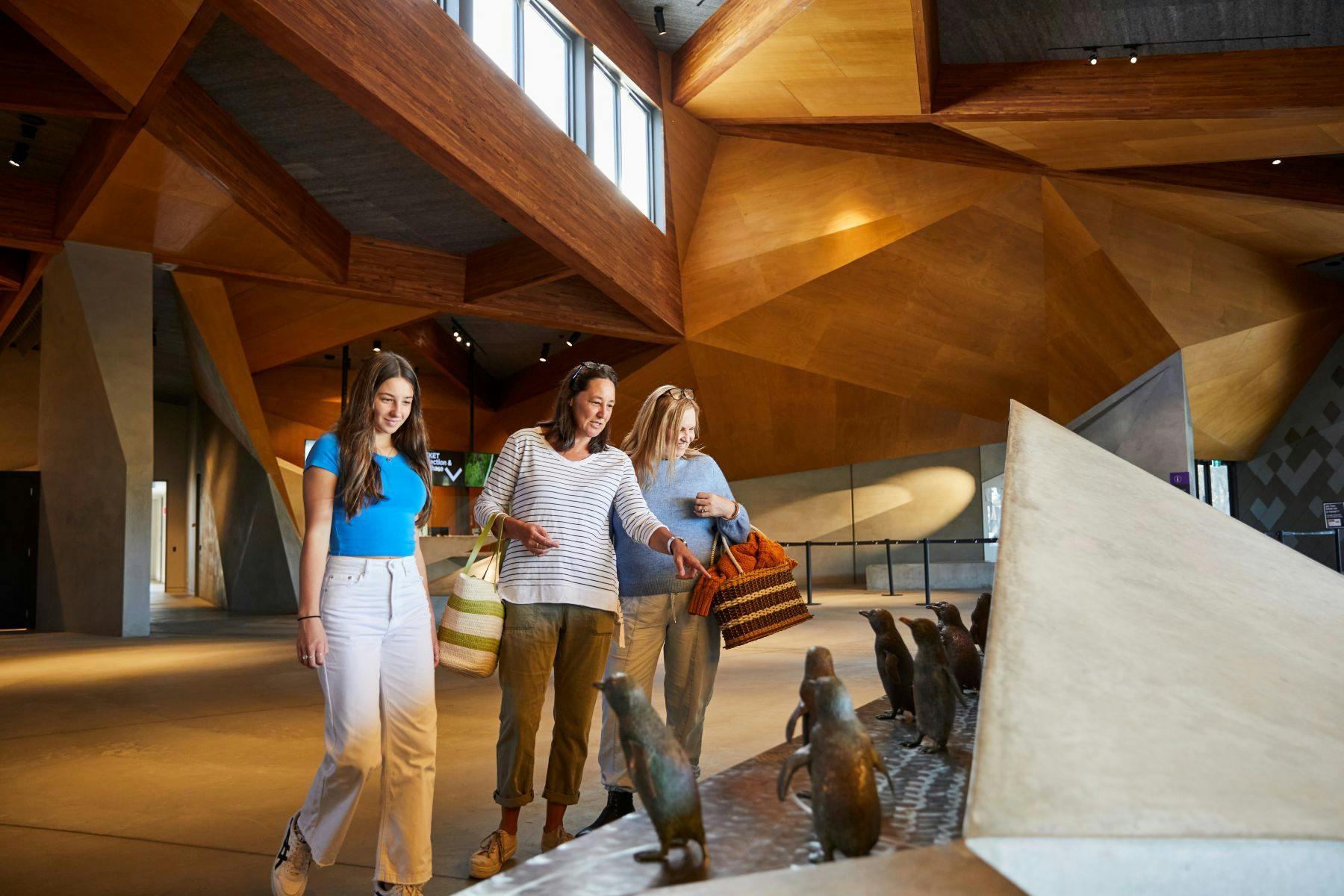 三位女士，每个人都拿着袋子，在现代木制建筑内欣赏企鹅雕塑。